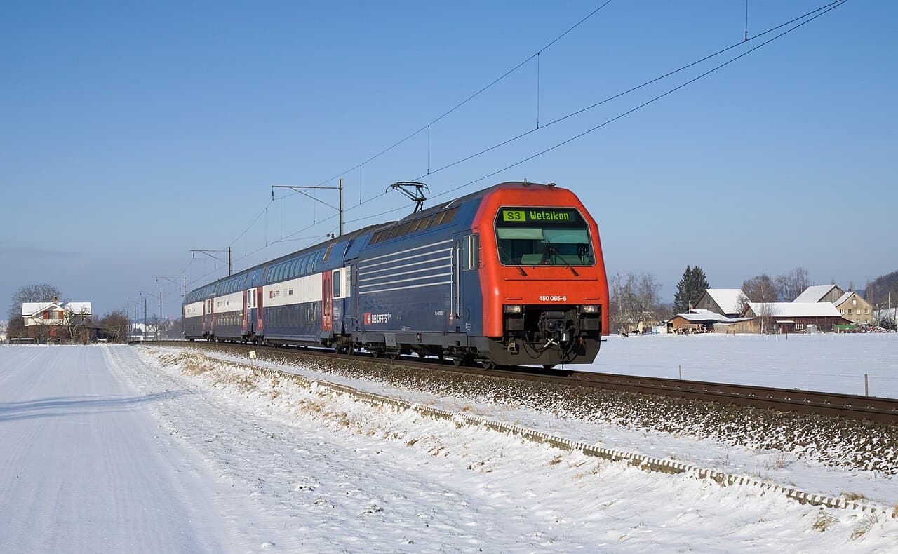 Blue Re 450 locomotive with red front pushing double-deck S-Bahn coaches through a snowy landscape