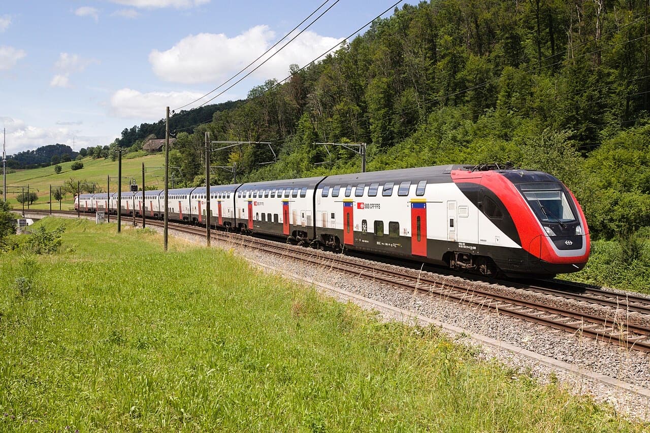 SBB FV-Dosto double-deck train in red and white, seen from the front three-quarter, running through green countryside near Tecknau